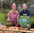 Alan Titchmarsh and Camilla Basset Smith posing outside in front of a table with various planters and strawberries on it. Behind them is the Haxnicks Vigoroot Balcony Planter.